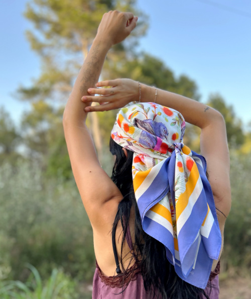 Woman seen from behind with a printed silk scarf in vibrant colours, violet and mustard, worn bandana-style over loose hair.